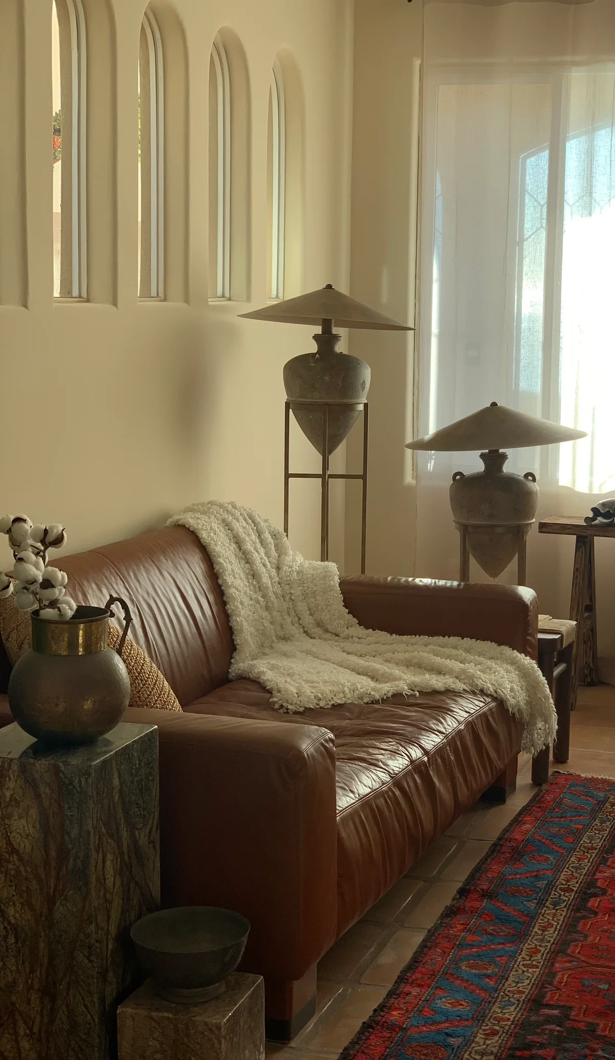 A long brown leather sofa under a sheepskin against a wall of tall arched niches, a pair of conical metal lamps either side of it and a heavy earthenware pitcher on a stone plinth in the foreground, on a red patterned rug.