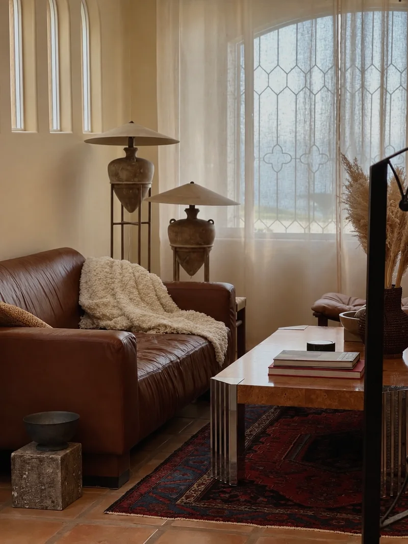 A tan leather sofa with a cream sheepskin throw over one arm, below a row of narrow arched windows and beside a leaded window hung with a sheer curtain, two conical metal lamps on slender stands behind it and a burr-figured low table on fluted steel legs holding books, on a red patterned rug over terracotta tiles.