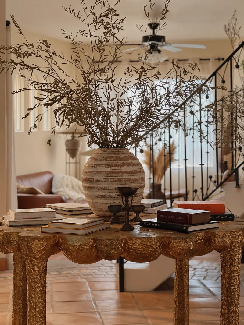 A tall striped terracotta vessel of dried olive branches on a scallop-edged gilt console, stacked books and a pair of dark candlesticks along the top, with a wrought-iron stair rail behind and the living room beyond.