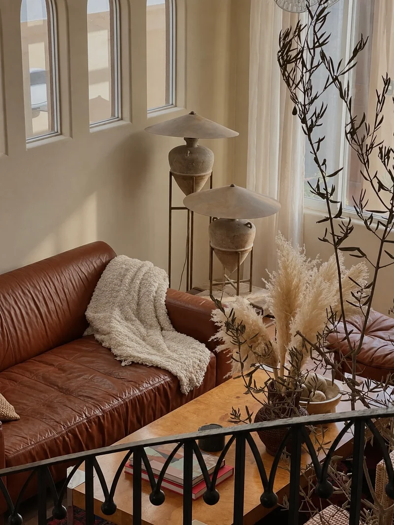 The tan leather sofa and its sheepskin seen past olive branches and pampas, the two conical lamps standing against a sheer curtain and the arched windows, and books stacked on the burr-figured table beyond a wrought-iron rail.