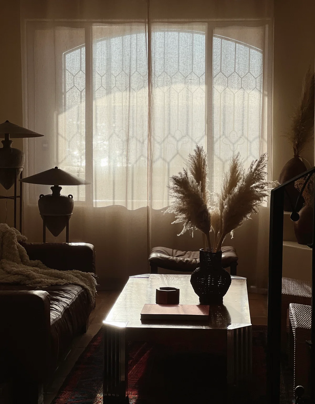 The same room in late light, the curtains lit from behind, a lamp and the pampas in silhouette over the coffee table.