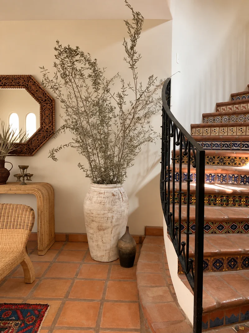 An octagonal wood-framed mirror on a white wall, a tall woven-textured urn of dried branches on the terracotta floor beside it, and the tiled stair rising at the right.