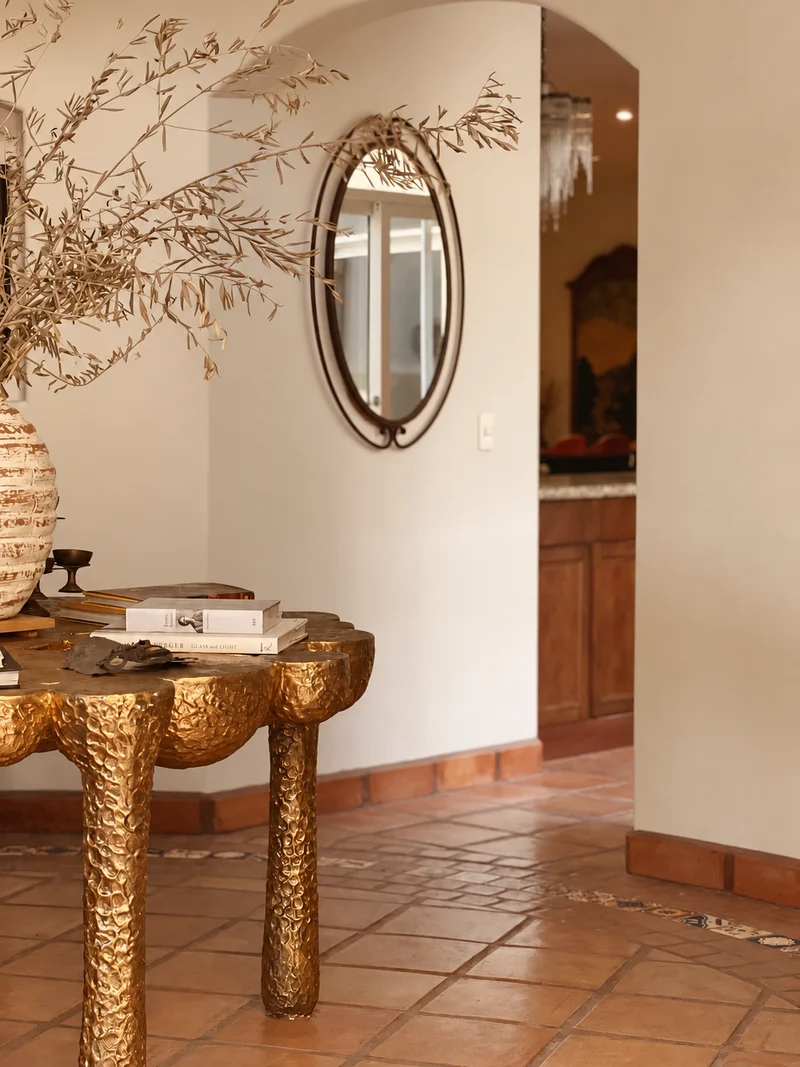 An oval mirror in a slim frame above the gilt console, dried branches arching out of a ribbed ivory pot, and a doorway through to the kitchen beyond.