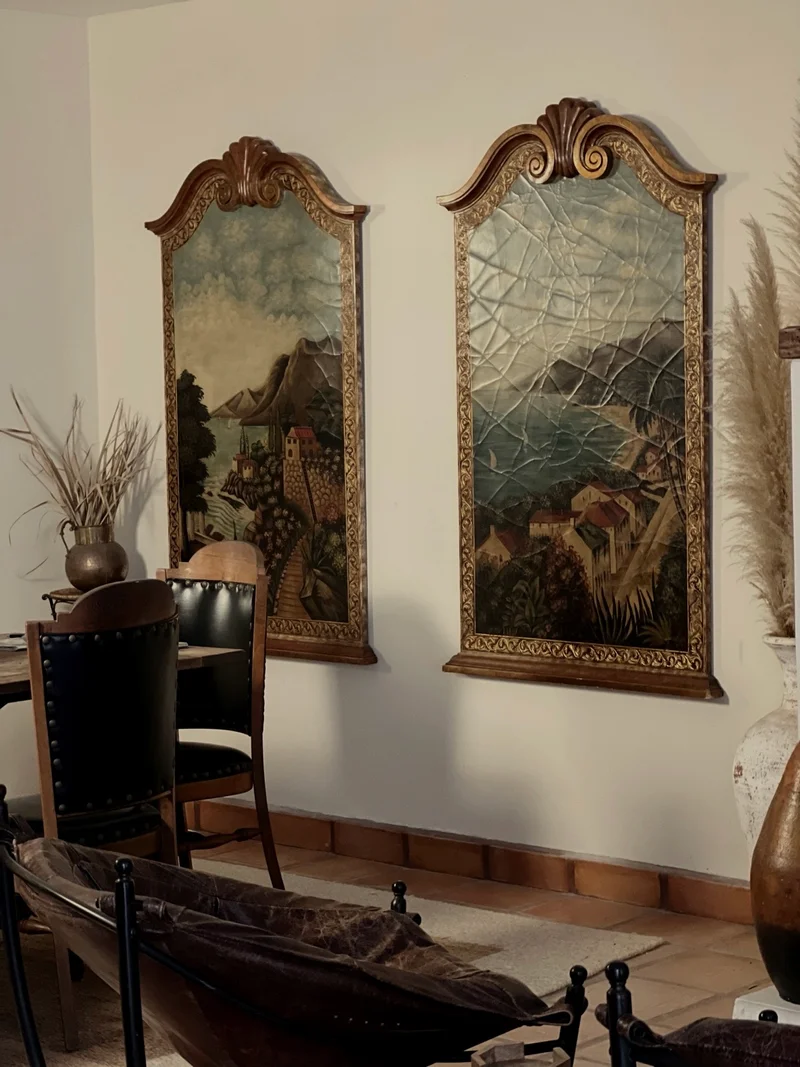 Two tall arched panels painted with hillside villages above a bay, in carved gilt frames with scrolled crests, hung on a plaster wall above a tiled skirting, with leather-backed chairs at a table and dried grasses in a metal pitcher to the left.