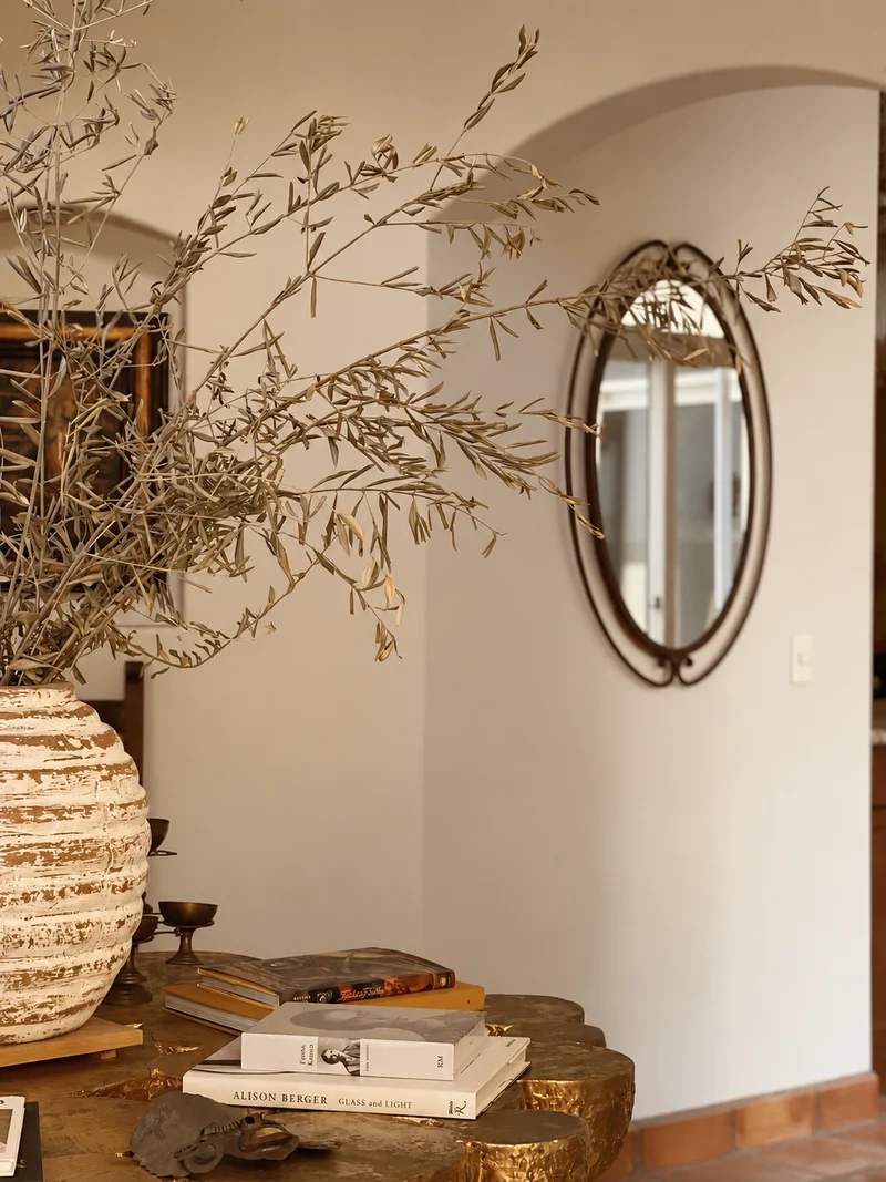 Tall dried olive branches arching out of a ribbed cream vessel standing on a gilt table stacked with books, an oval iron-framed mirror on the plaster wall beyond reflecting a window, and an arched opening through to the terracotta-tiled floor at the right.