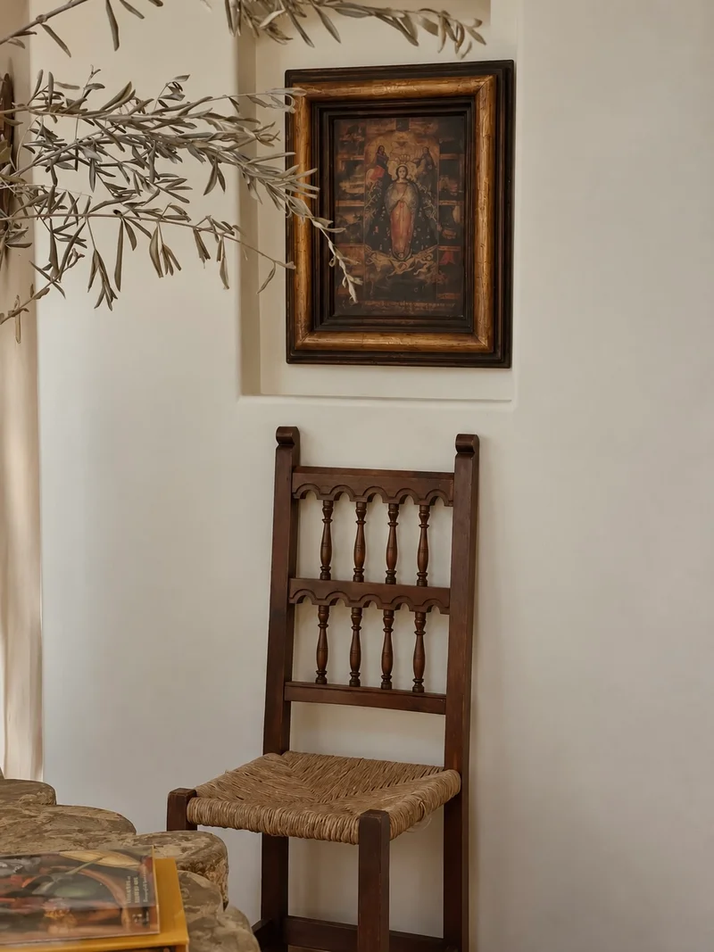 A spindle-backed timber chair with a woven rush seat standing against a plaster wall, a small dark-framed painting set into a shallow niche above it, and olive branches reaching in from the left.