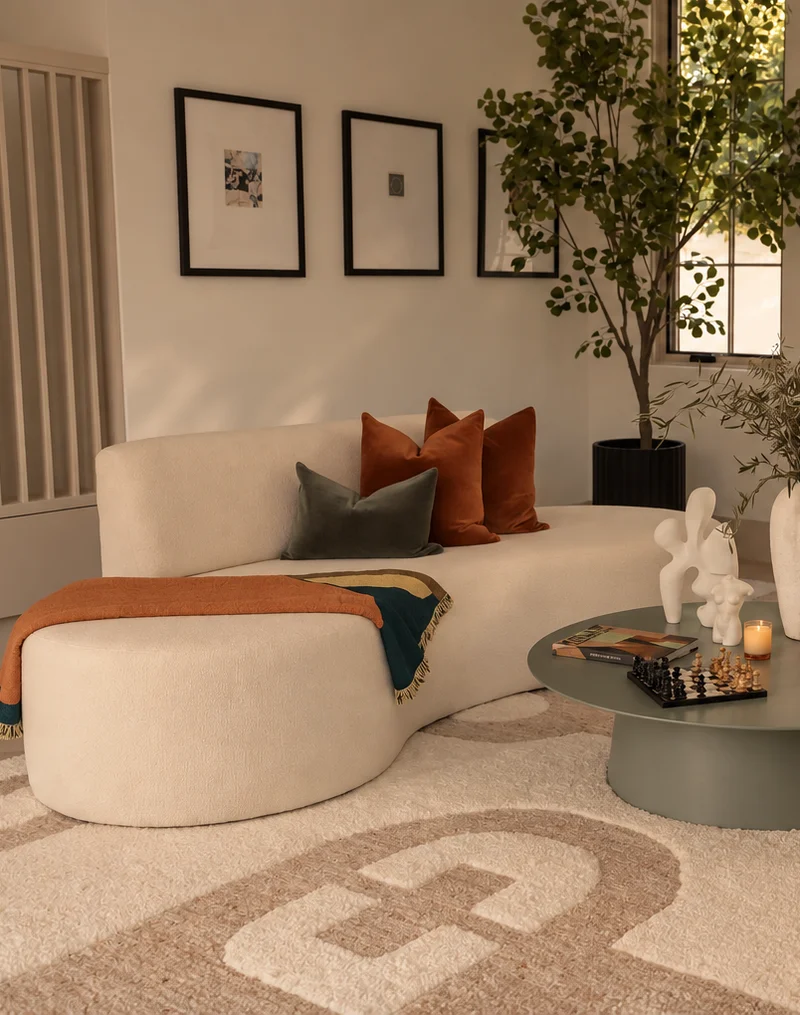 A curved cream boucle sofa and matching ottoman on a patterned wool rug, rust and sage cushions along the back and a striped throw over the arm, with three framed prints above and a sage-green round table holding a chess set, books and a lit candle beside a tall potted tree at a black-framed window.