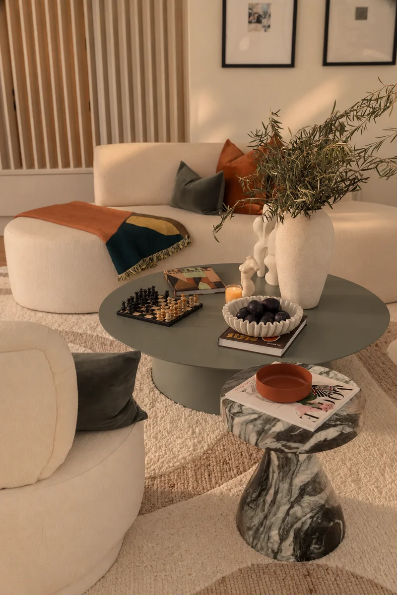 Olive branches in an ivory vase on the green table against the fluted timber screen, the framed prints beyond, and the marbled side table at the right.