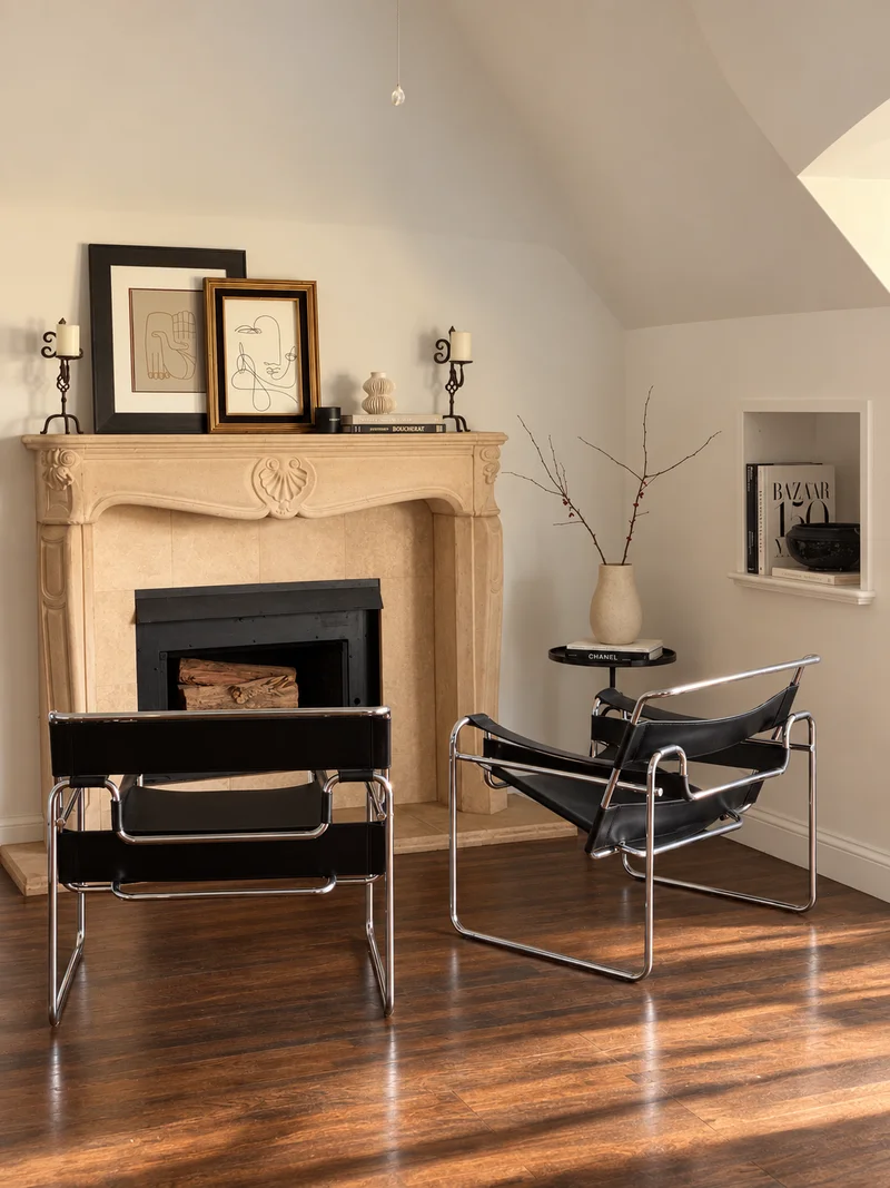 A carved limestone fireplace under a white vaulted ceiling, two black leather and chrome sling chairs facing it, and framed pictures propped along the mantel.