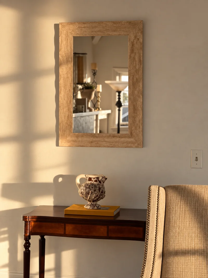 A limed-wood framed mirror above a dark wood console holding a painted ceramic pitcher and a yellow book, with a striped upholstered chair beside it.