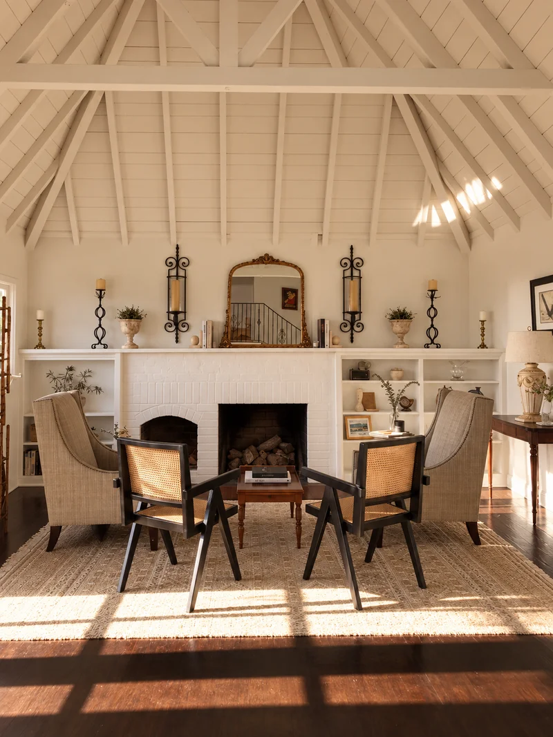 A white-beamed vaulted room, a fireplace flanked by built-in shelves under a gilt mirror and iron sconces, and cane-and-timber chairs drawn around a low table on a jute rug in low sunlight.