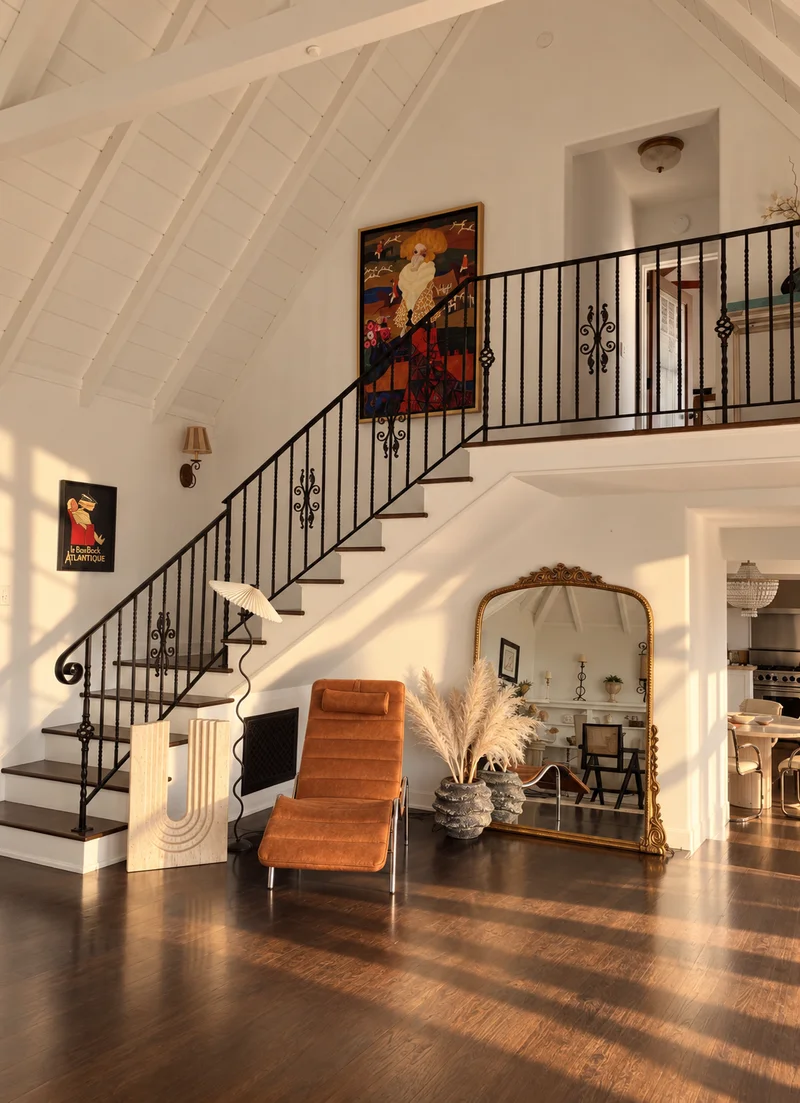 A double-height hall under white-painted rafters: an iron-railed staircase climbing to a gallery landing, a tan velvet lounge chair and a fluted plaster console below, pampas in a pair of stone urns, and a tall gilt mirror against the wall.