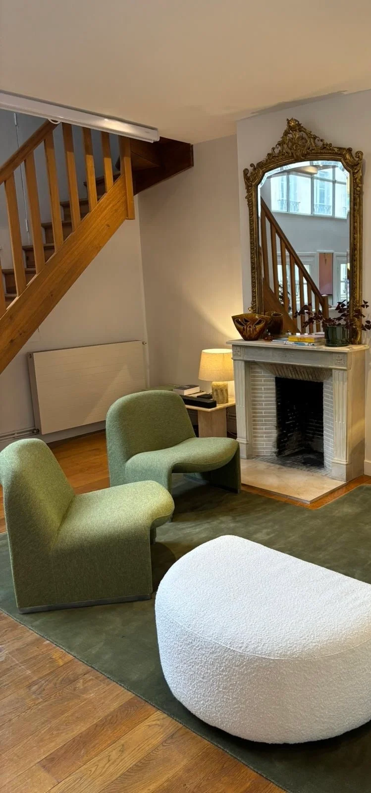 A Parisian sitting room: two green upholstered chairs and a cream boucle ottoman on a moss-green rug, a white fireplace under a tall gilt mirror, and an open-tread wooden stair climbing the wall behind them.