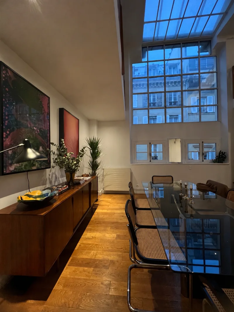 A long walnut sideboard along a white wall under a framed picture and a black task lamp, a dark glass dining table with cane-and-chrome chairs drawn up to it beyond, lit from a glazed roof and the tall windows behind.