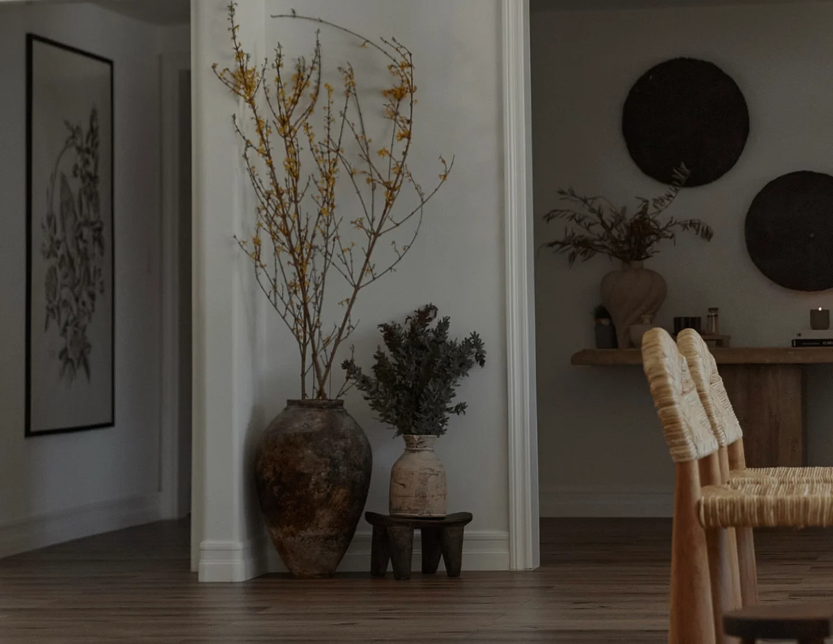 A framed botanical drawing on a plaster wall, a tall terracotta urn of dried branches at the corner, round woven discs hung above a console, and pale woven chairs drawn up to it.