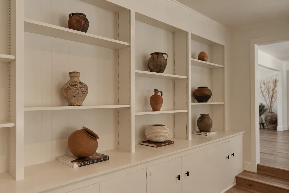 A wall of built-in cream shelving holding a collection of antique terracotta and stoneware pots, each on its own shelf and several standing on books, with an opening through to a further room beyond.