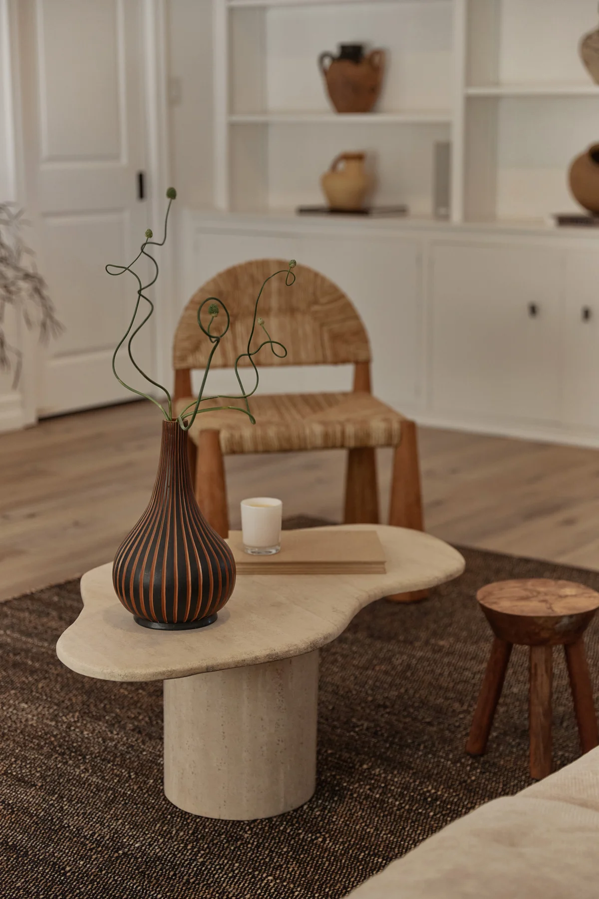 A curved travertine coffee table on a dark woven rug, holding a striped gourd-shaped vase of wiry branches and a candle, with a small round timber stool beside it and a woven-back chair and pale shelving beyond.