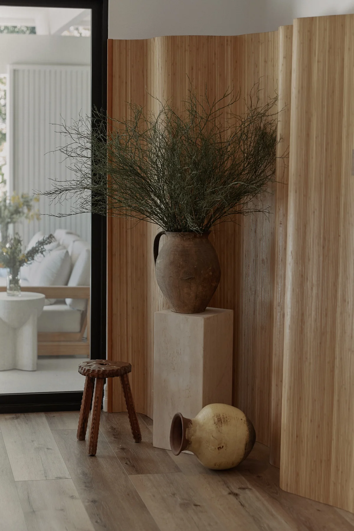An antique terracotta vessel of dried branches on a travertine pedestal, against a fluted timber screen, with a carved wooden stool and a pale glazed jar on the oak floor beside it.