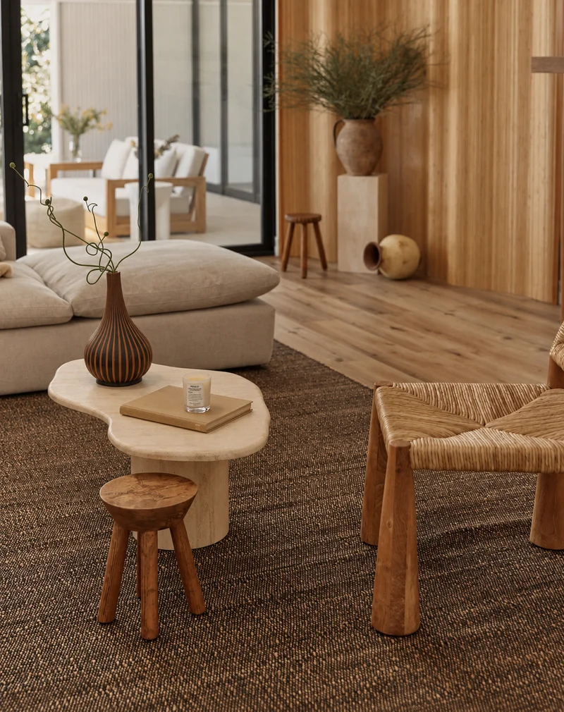 The kidney-shaped stone table from the other side, the ribbed dark vase and a candle on it and a small timber stool tucked beneath, with a sectional sofa to the left, a woven chair to the right, and glazed doors onto a furnished terrace past a timber-clad wall.