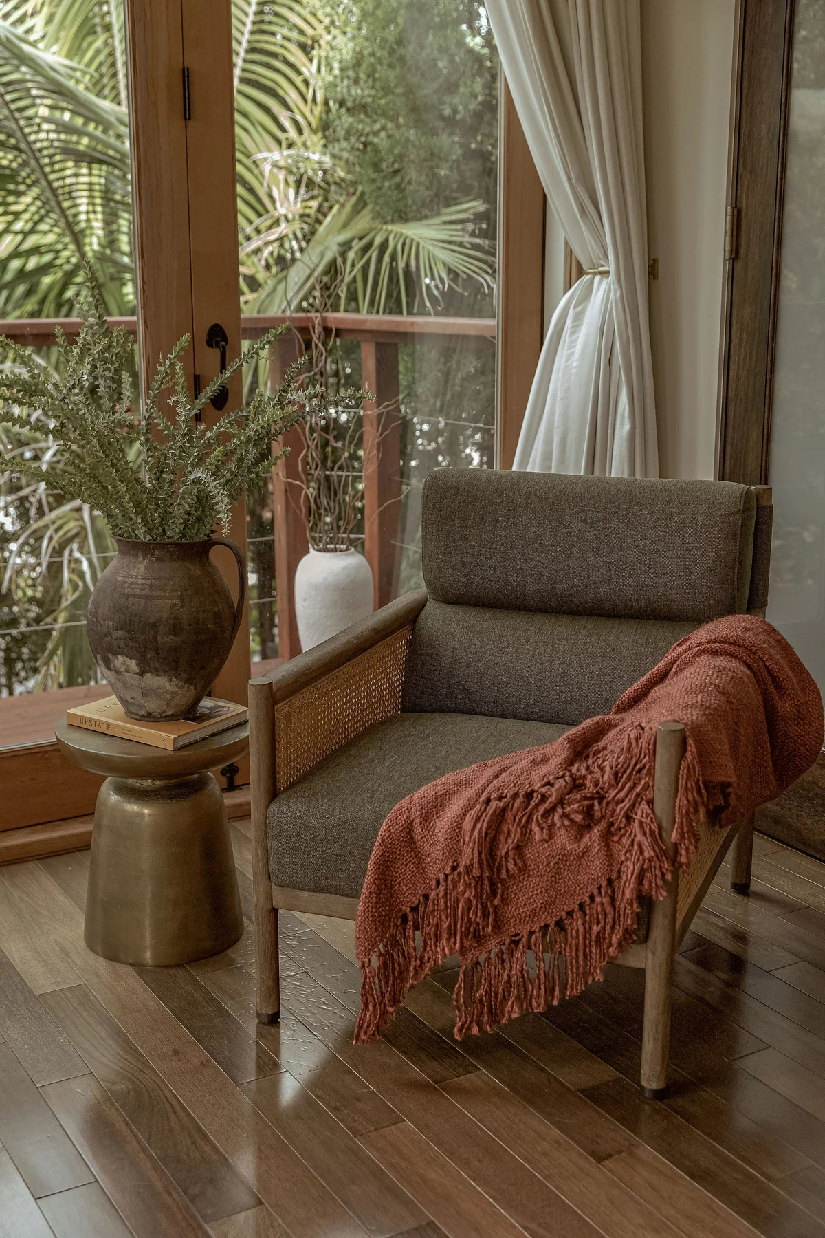 A grey upholstered armchair with a rust fringed throw over one arm, a brass drum table beside it holding books, and a rustic pitcher of dried grass by the sliding doors onto the bamboo.