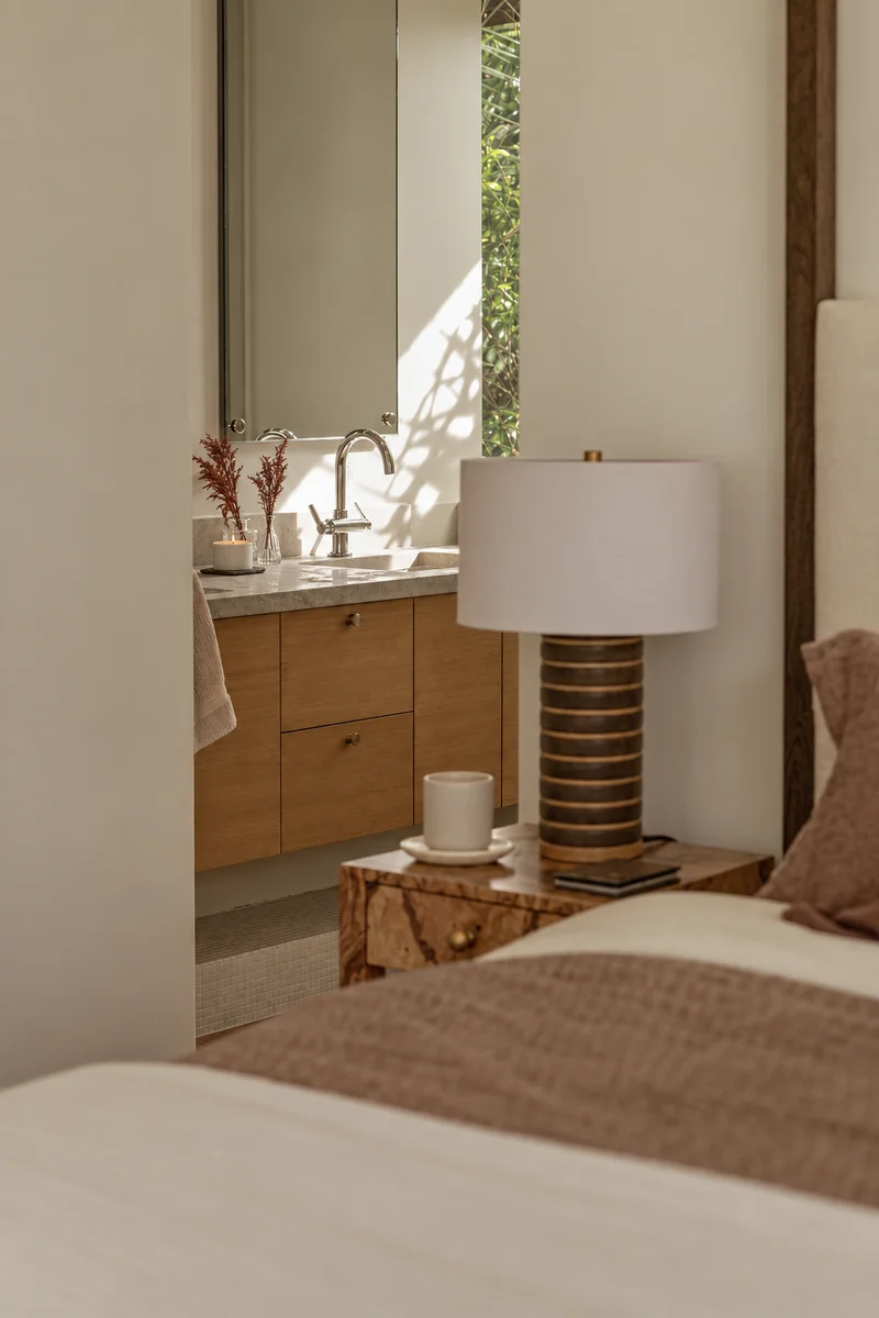Looking past the bed and its lamp into the bathroom: an oak vanity under a stone top and a tall mirror, a candle and dried rust-coloured stems beside the basin, and a slot window onto greenery throwing branch shadows across the wall.