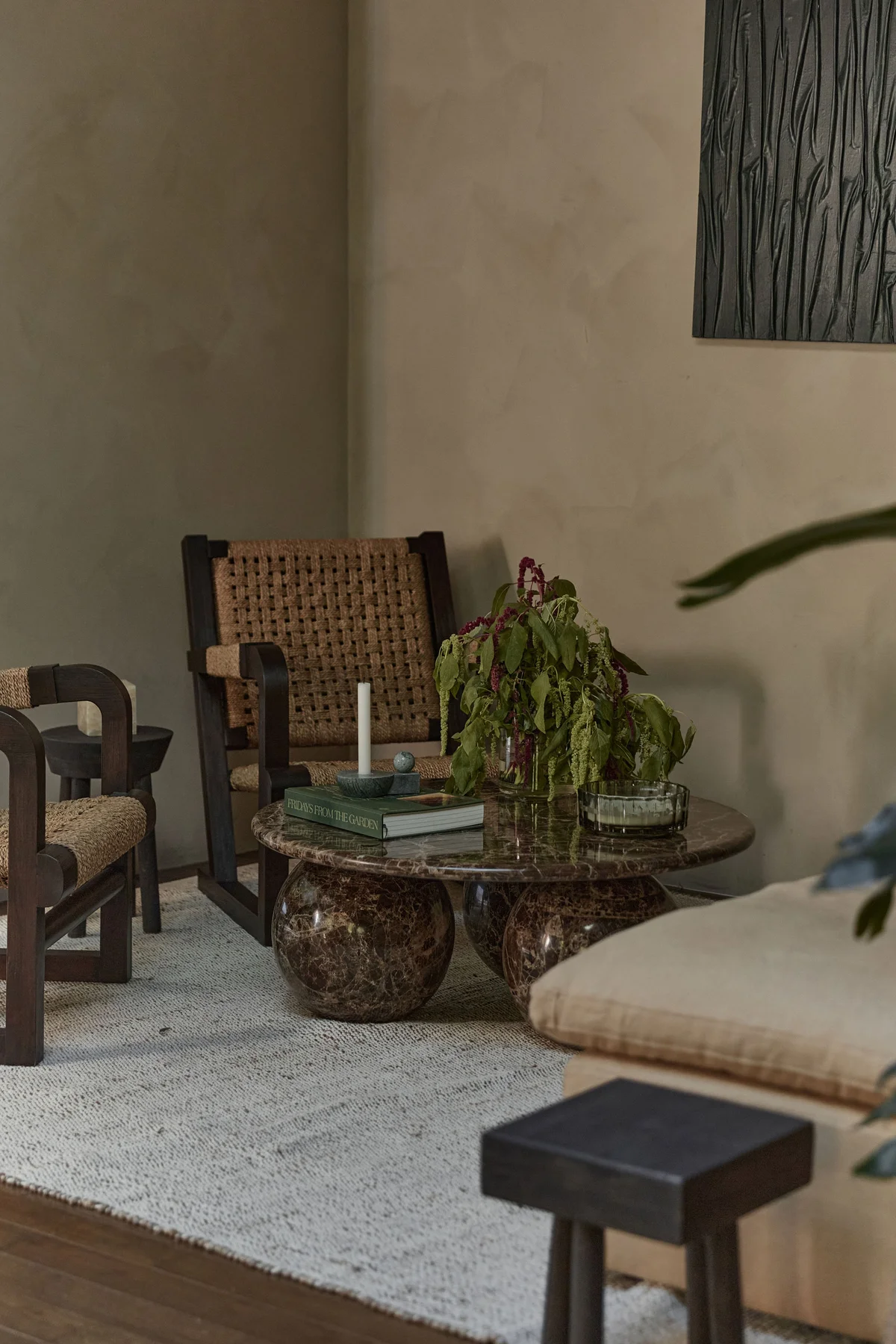 Two low timber-and-cane armchairs facing a round stone coffee table across a cream rug, a trailing plant and books on the table, a long ottoman at the right and a textured plaster wall behind.