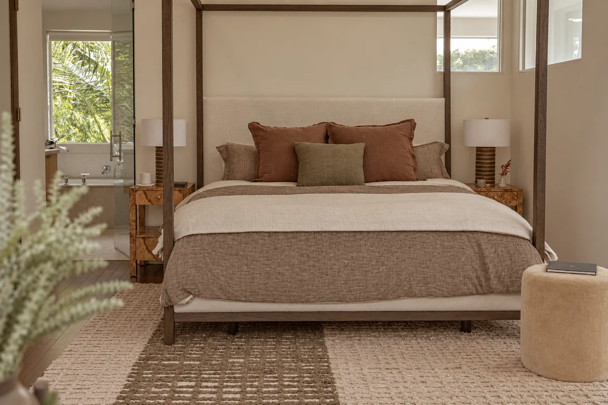A four-poster bed with a tall upholstered headboard, rust and olive cushions along it and a knitted throw across the foot, burr-figured nightstands and ribbed lamps either side, layered jute rugs underfoot and a bathroom visible through the opening beyond.