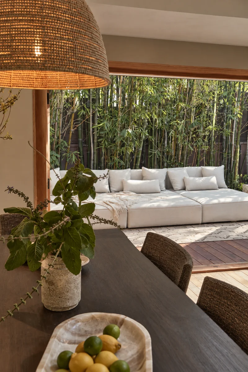 A leafy plant in a mottled stone pot and a wooden bowl of lemons and limes on the dark dining table, the wide woven pendant above, and past the folded-back doors a long cushioned daybed on the deck with bamboo close behind it.