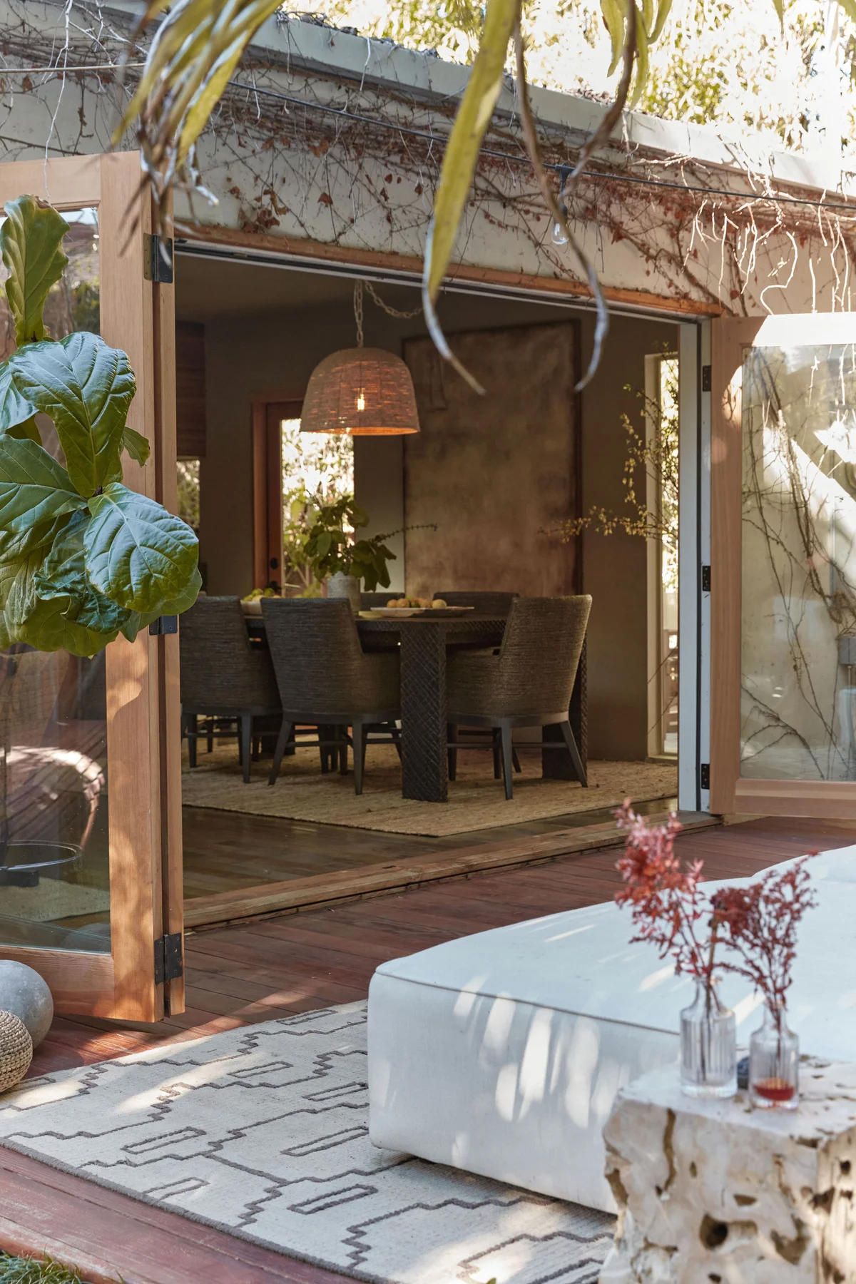 The dining table seen from the deck through folded-back doors, a fiddle-leaf fig at the threshold and a low white daybed on the patterned rug outside.