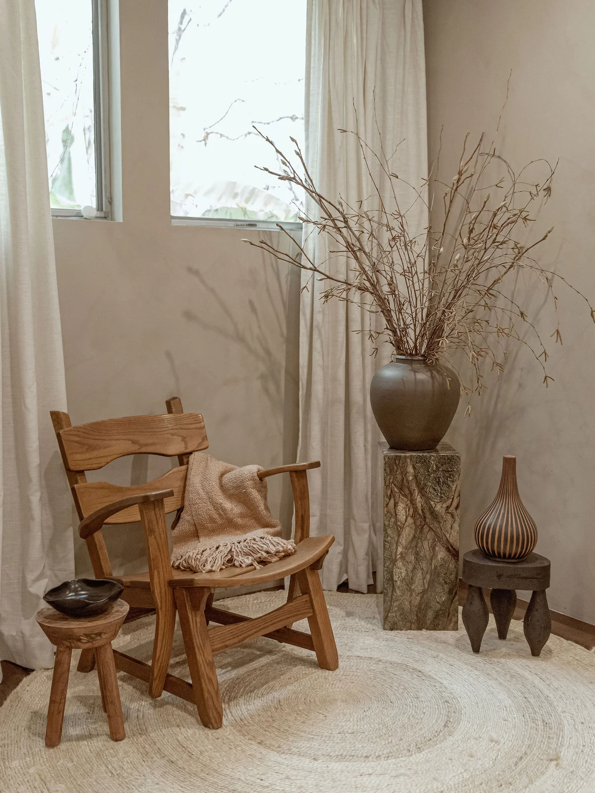 A timber lounge chair with a cream throw on a round jute rug, a three-legged stool with a dark bowl in front of it, and a tall dark vessel of branches on a stone pedestal against ivory curtains.