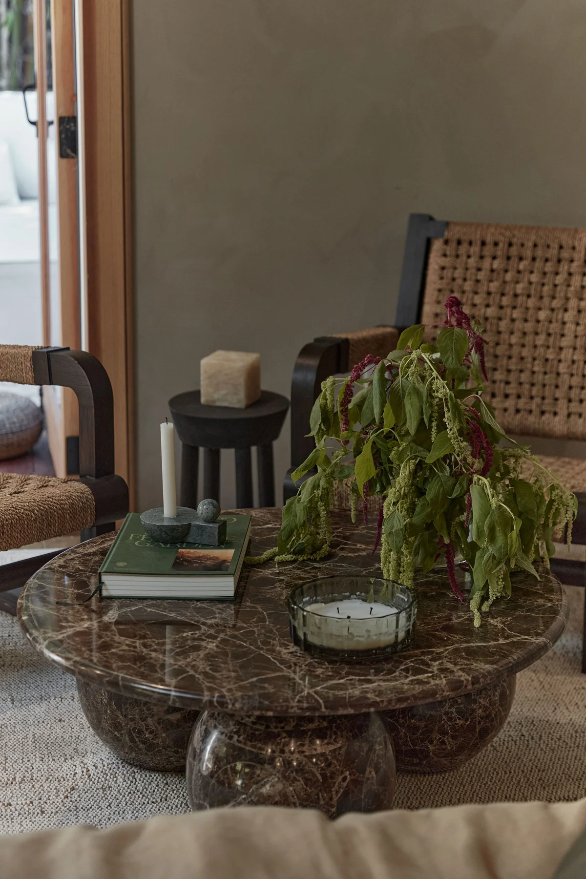 The stone table close up — a trailing plant, a green candle in a glass and a stack of books on the dark veined top, with the cane chair backs behind and daylight through an open door.