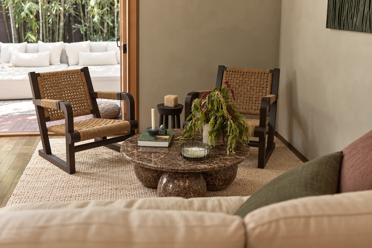 Two dark-framed lounge chairs with woven rope backs and seats drawn up to a round marbled coffee table on ball feet, a candle and books on it beside a trailing plant, a pale sofa in the foreground and folded-back doors onto a cushioned daybed on the deck beyond.
