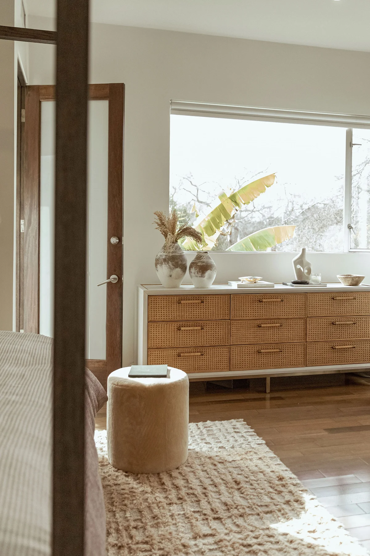 A cane-fronted sideboard set with pale vessels, seen past a door frame, with a white sofa and a round stone side table on a striped rug in the foreground.