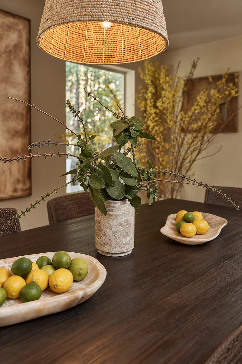 Looking along the dark timber dining table: a mottled stone vase of leafy branches and flowering stems, a shallow banded-stone dish of lemons and limes beyond it and a larger one nearer, under the wide woven pendant, with tall yellow blossom in the corner of the room.